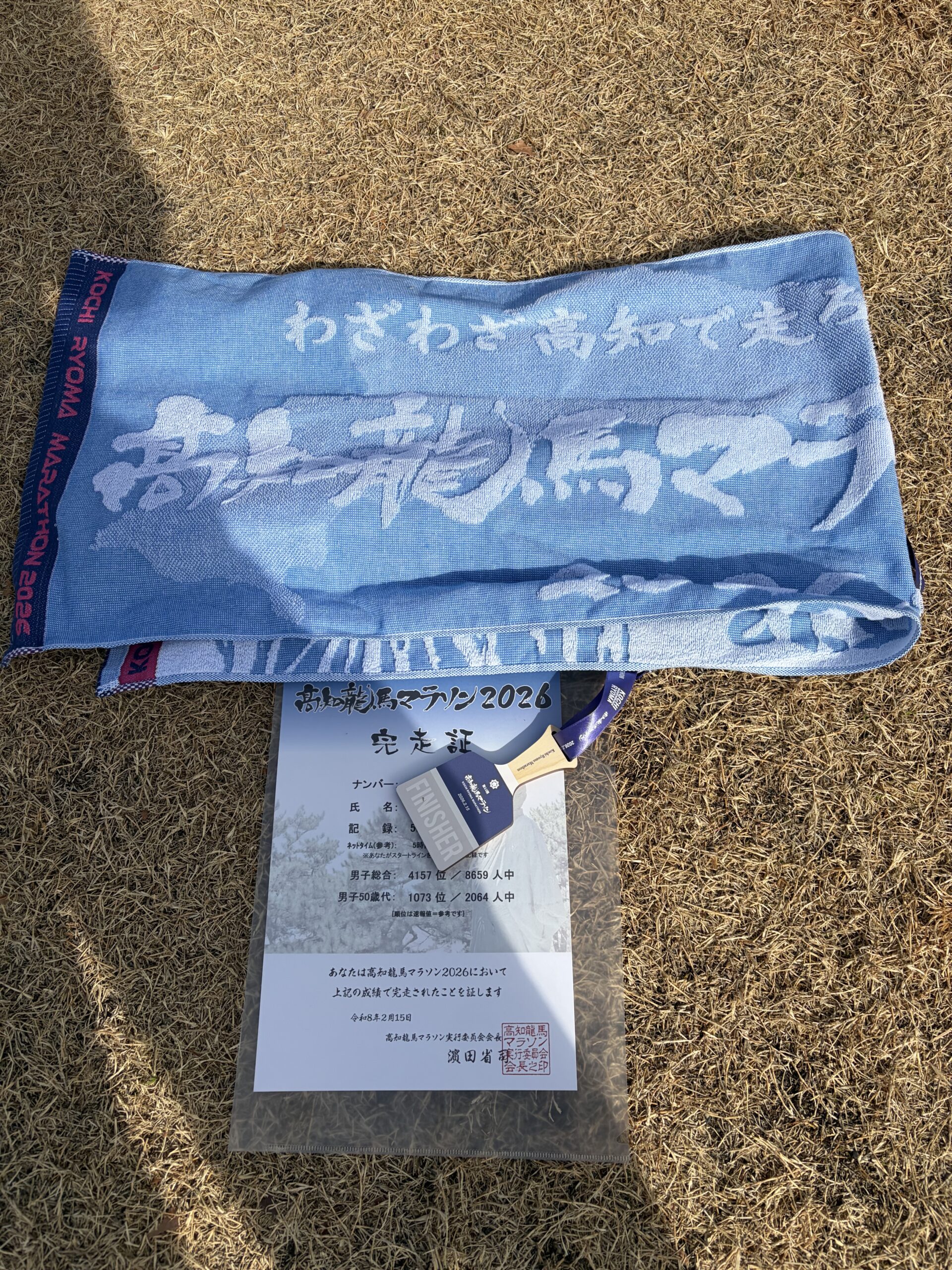 芝生の上に置かれた2026年高知龍馬マラソンの完走証、水色のフィニッシャータオル、そして「FINISHER」と刻印された木製の鳴子メダル。